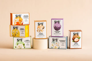Seven colorful SOF soap boxes with floral and fruit illustrations are arranged in a neat display on a beige background. Varieties include Orange Blossom, Lemon Verbena, Sweet Almond, Lavender Fields, Lush Gardenia, Blooming Jasmine, and Shea Butter.