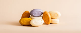 Nine colorful oval soap bars, mostly in shades of beige, orange, and purple, are arranged in a small pile on a light beige background. The word “SOF” is embossed on each bar.