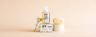 A bottle of SOF Blooming Jasmine foaming hand soap and a bar of soap are displayed with their packaging, a small sprig of flowers, and a beige background.