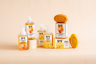 A display of SOF brand soap products, including liquid hand soaps in pump bottles and bar soaps with packaging, featuring scents like Lemon Verbena and Orange Blossom Honey on a neutral beige background.