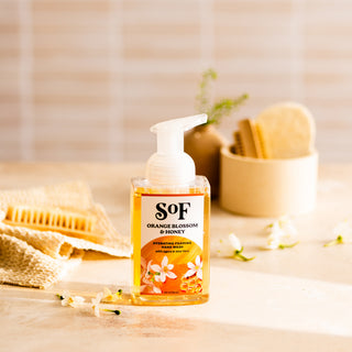 A bottle of Orange Blossom & Honey Hydrating Foaming Hand Wash—phthalate free—rests on a light surface, surrounded by white flowers, a textured towel, a brush, and bath accessories in softly lit bathroom decor.