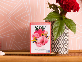 A box of Wild Rose Moisturizing Bar Soap, made with natural colorants and SLS free, sits on a bamboo tray beside a vase with red flowers and green leaves, against a pink patterned wall.