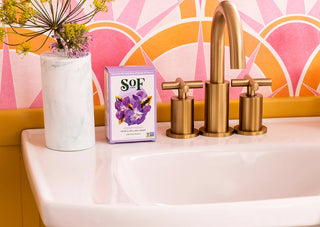 A box of Fresh Violet Moisturizing Bar Soap with purple flowers rests on a white sink near a vase of yellow flowers and brass faucet handles, set against a pink and orange geometric patterned wall.