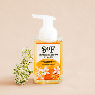 A bottle of Orange Blossom & Honey Hydrating Foaming Hand Wash with a pump and orange and white floral design sits beside a sprig of white flowers on a neutral beige background.