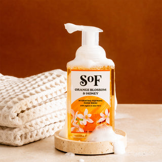 A bottle of Orange Blossom & Honey Hydrating Foaming Hand Wash, SLS/SLES free, rests on a bubble-covered round dish with white towels in the background against a brown backdrop.