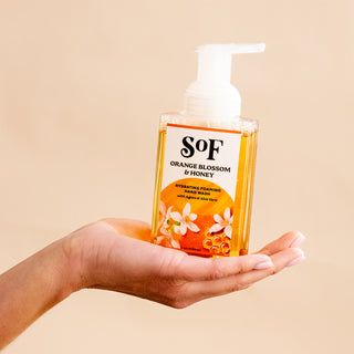 A hand holds a clear bottle of Orange Blossom & Honey Foaming Hand Soap from the SoF essentials line, featuring a pump dispenser, set against a beige background—perfect for your daily cleanse with its beautifully layered scents.