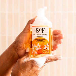 A person with soapy hands holds Orange Blossom & Honey Hydrating Foaming Hand Wash—free from parabens and phthalates—against a light tiled background.