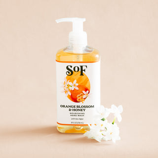 A bottle of Orange Blossom & Honey Nourishing Hand Wash with a pump and floral label sits beside white flowers on a beige background. This nourishing hand wash is paraben free.