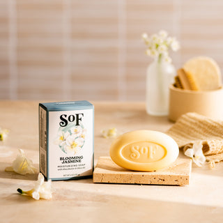 A Blooming Jasmine Moisturizing Bar Soap rests on a cork tray beside its floral box, with white jasmine flowers, a towel, and bath accessories softly blurred in the background.