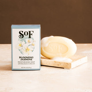 A box of Blooming Jasmine Moisturizing Bar Soap stands next to an oval bar with rich lather and bubbles, both arranged on a beige stone surface against a brown background.