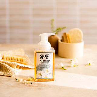 A bottle of Blooming Jasmine Hydrating Foaming Hand Wash, SLS and paraben free, sits on a beige surface with jasmine flowers, a honeycomb, and bath brushes in a softly lit, inviting setting.