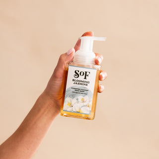 A hand holds a clear bottle of Blooming Jasmine Hydrating Foaming Hand Wash with a pump dispenser against a beige background. The label shows white jasmine flowers and highlights the hydrating, foaming, paraben-free formula.