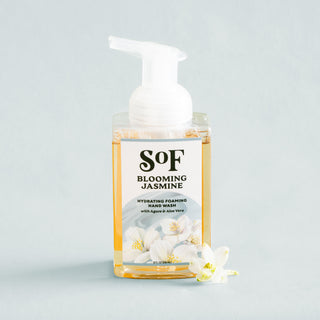 A bottle of Blooming Jasmine Hydrating Foaming Hand Wash, SLS- and paraben-free with agave and aloe vera, features a white pump and floral label, set on a light surface with white jasmine flowers beside it.