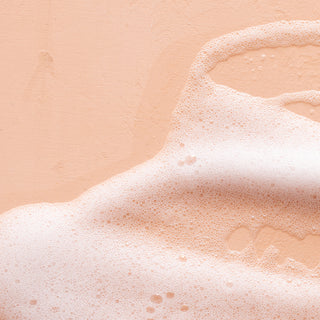Close-up of white, foamy bubbles from Lemon Verbena Hydrating Foaming Hand Wash spread over a peach surface, creating a textured, pastel-toned backdrop.