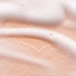 A close-up of frothy, peach-hued liquid from the Lemon Verbena Bundle reveals a patch of exposed surface rimmed with bubbles and foam, suggesting a refreshing daily cleanse.