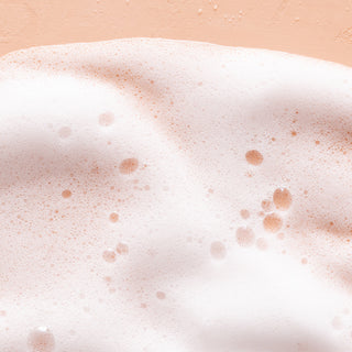 A close-up of white soap bubbles and foam from Sweet Almond Hydrating Foaming Hand Wash covers a light peach surface, with various bubble sizes highlighting its soft, frothy texture and deeply hydrating formula.