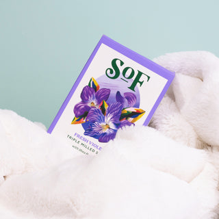 A Fresh Violet Moisturizing Bar Soap with a purple violet flower on the packaging rests on a soft, white blanket against a pale blue background.