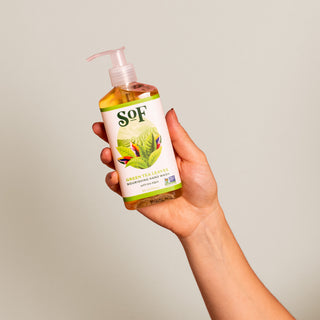 A hand holds a clear pump bottle labeled “Green Tea Leaves Nourishing Hand Wash,” adorned with green tea leaves and a bird. This moisturizing wash, enriched with essential oils, is shown against a simple light background.
