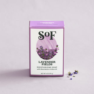 A box of Lavender Fields Moisturizing Bar Soap, enriched with shea butter and olive oil, sits on a light purple background. The packaging features lavender flowers and purple accents, with lavender buds scattered nearby.