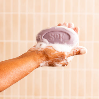 A hand holds the Lavender Fields Moisturizing Bar Soap, a purple, triple-milled bar embossed with "SOF," covered in white lather against a beige tiled background.