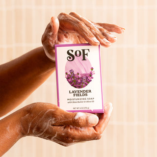 With soapy hands, a person holds a box of the Lavender Fields Bundle moisturizing soap. The white and purple box shows lavender flowers. Light-colored tiles are in the background.