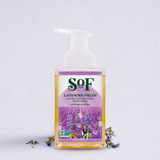 A clear bottle of Lavender Fields Hydrating Foaming Hand Wash with a pump sits on a light surface. The label shows lavender flowers and text, while sprigs of lavender and soothing aloe vera are placed nearby.