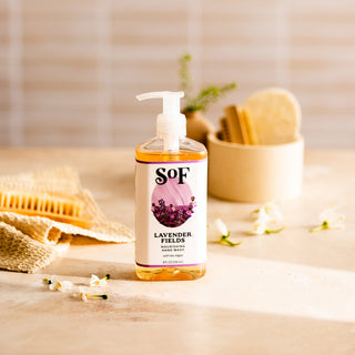 A bottle of Lavender Fields Nourishing Hand Wash with a pump sits on a light surface, surrounded by natural spa items and white flowers. The softly blurred beige background enhances the calm, fresh atmosphere.