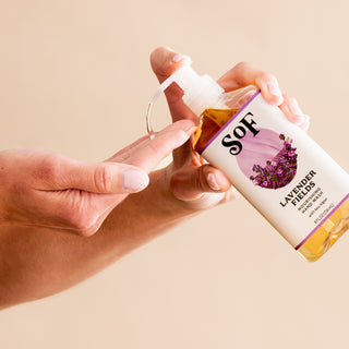 A hand presses the pump of a Lavender Fields Nourishing Hand Wash bottle, dispensing SLS-free soap onto another hand. The label features a lavender blossom illustration against a plain, neutral background.