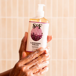 Soapy hands hold a clear pump bottle of Lavender Fields Nourishing Hand Wash, which is paraben free, against light tiles. The label shows purple lavender flowers with a lavender-colored design.