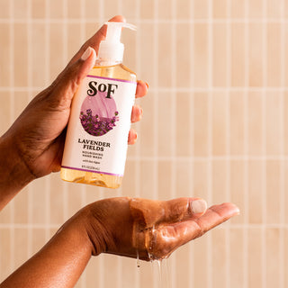 A person dispenses Lavender Fields Nourishing Hand Wash—SLS and paraben free—onto their palm from a pump bottle against a beige tiled background.