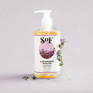 A bottle of Lavender Fields Nourishing Hand Wash sits on a light purple surface next to fresh lavender. The label shows blooming lavender flowers, and the hand wash comes with a pump top and is paraben free.