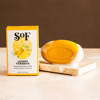 A yellow box labeled Lemon Verbena Moisturizing Bar Soap stands beside a yellow oval soap embossed with “SoF,” both resting on a stone surface with lather against a brown background.
