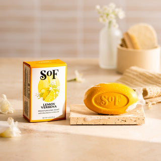 A yellow bar of Lemon Verbena Foaming Hand Soap and Bar Soap rests on a stone dish beside its box, with blurred bath accessories and a vase of white flowers in the background—perfect for your daily cleanse.