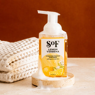 A bottle of Lemon Verbena Hydrating Foaming Hand Wash sits on a round stone tray with foam at its base, paired with stacked textured towels against a warm brown background. This formula is paraben free and SLS free.