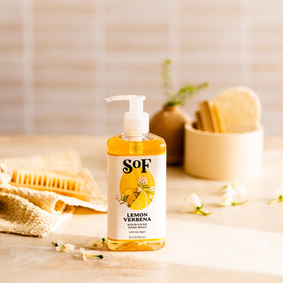 A bottle of Lemon Verbena Nourishing Hand Wash, SLS and paraben free, sits on a light countertop surrounded by flowers, a textured cloth, a wooden brush, and a bowl of sponges in soft, neutral lighting.