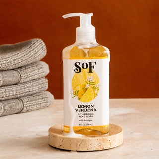 A bottle of Lemon Verbena Nourishing Hand Wash with a pump sits on a stone coaster beside neatly folded gray towels against a brown backdrop.