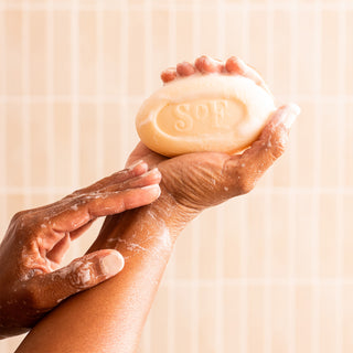 A person is lathering their arm with a Lush Gardenia Moisturizing Bar Soap, engraved with SOF, against a tiled background.