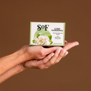 A person holds a green box of Lush Gardenia Moisturizing Bar Soap, enriched with organic shea butter and olive oil, featuring a white flower illustration, against a plain brown background.