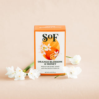 An upright box of Orange Blossom & Honey Moisturizing Bar Soap is shown surrounded by white flowers on a beige background, with packaging decorated with orange and floral designs.