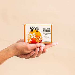 A person holds a box of Orange Blossom & Honey Moisturizing Bar Soap, with orange slices, honeycomb, and white blossoms on the packaging, set against a plain beige background.