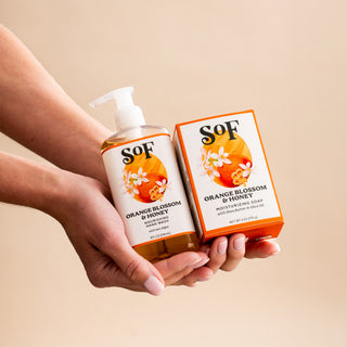 Two hands holding a bottle of SOF Orange Blossom & Honey moisturizing hand pump soap and a matching box with floral artwork on a beige background.