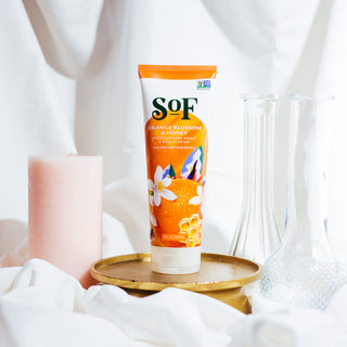 An orange tube of Orange Blossom & Honey Hand and Body Cream sits on a gold tray, with a pink candle, two clear glass vases, and white fabric in the background.
