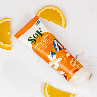 A tube of Orange Blossom & Honey Hand and Body Cream rests on a white surface, surrounded by three orange slices. The tube features orange, honeycomb, and floral designs.