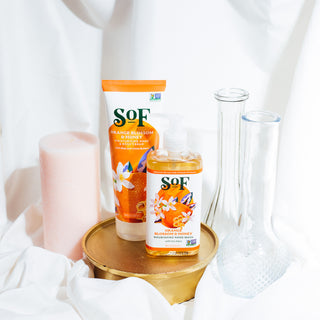 The Orange Blossom & Honey Liquid Hand Wash and Hand & Body Cream are displayed on a round gold tray with a pink candle and two clear glass vases, all set against a white fabric background.