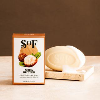 A box of Shea Butter Moisturizing Bar Soap stands next to an oval triple-milled bar stamped with the SoF logo, resting on a beige stone dish. The neutral brown background highlights its organic shea butter and rich lather.