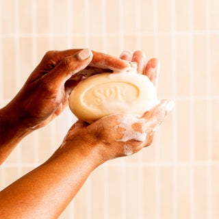 Two hands lather a Shea Butter Moisturizing Bar Soap engraved with "SOF" against a backdrop of light-colored tiles.
