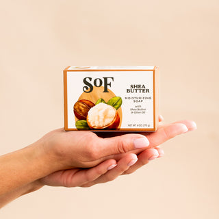 A hand holds a rectangular box of Shea Butter Moisturizing Bar Soap with organic shea butter and olive oil, set against a plain beige background. The triple-milled soap packaging displays an image of shea nuts.