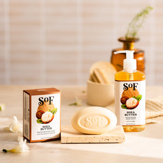 A box, bar, and pump bottle of Shea Butter Liquid Hand Soap and Bar Soap are displayed on a beige surface with white flowers and bathroom accessories. The soap bar, embossed with the SOF logo, gently cleanses your skin.