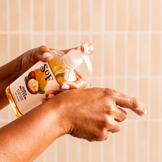 A person dispenses Shea Butter Nourishing Hand Wash from a clear SOF bottle onto the back of their hand, with a light tiled background.