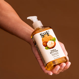 A hand holds a clear pump bottle of Shea Butter Nourishing Hand Wash against a solid brown background. The label shows shea nuts and leaves, highlighting an SLS/SLES-free formula for gentle, safe cleansing.